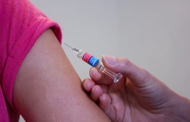 Une personne s'aprète a vacciner. Un bras et découvert, la personne dont on ne vois que le bras, porte un chandail rose, on ne voit que la main de la personne qui vaccine.