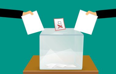 Deux mains (une de chaque côté) déposant leur vote politique dans une boite clair sur une table en bois. Cette image à un fond turquoise foncé.