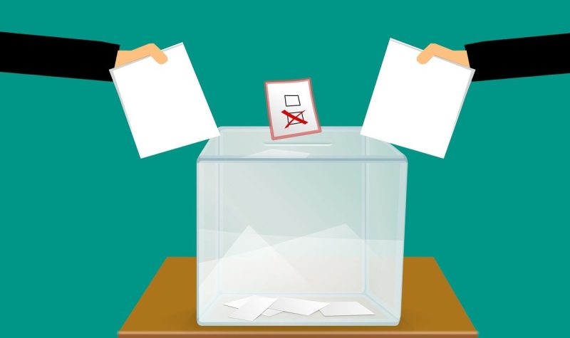 Deux mains (une de chaque côté) déposant leur vote politique dans une boite clair sur une table en bois. Cette image à un fond turquoise foncé.