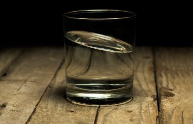 A glass filled with water sits on a wooden table. A dark room sits as the backdrop to the photo.