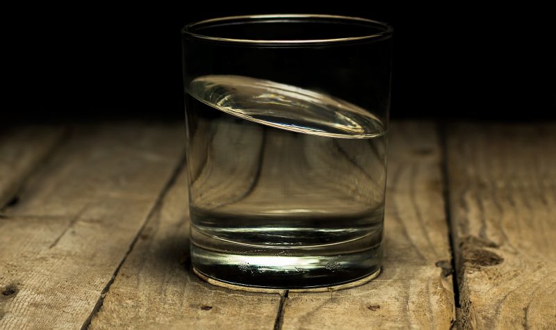A glass filled with water sits on a wooden table. A dark room sits as the backdrop to the photo.