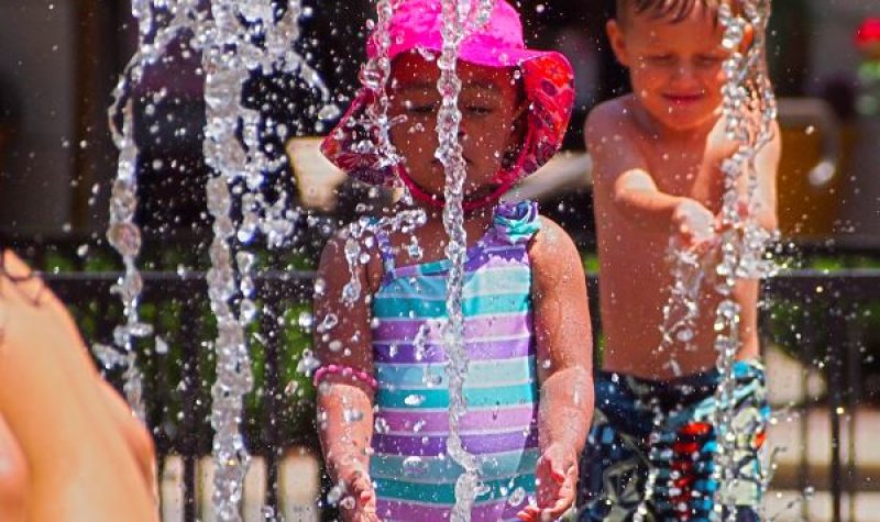 Children play in a water park. Water spouts up as a small girl in a striped pink purple and blue one piece swimsuit and a pink sun hat closely stares and plays. Behind her and on the right on the photo a shirtless bot in blue, white, black and grey Volcom swim trunks catches water from another spout.