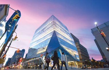 A large glass building in the middle of a street intersection with people walking infront.