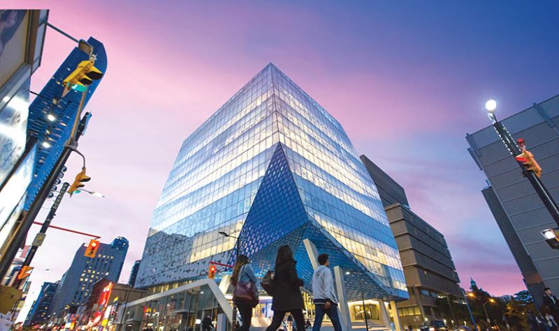 A large glass building in the middle of a street intersection with people walking infront.