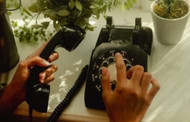 Hands dial an old fashioned phone on a white table with potted plants around it.