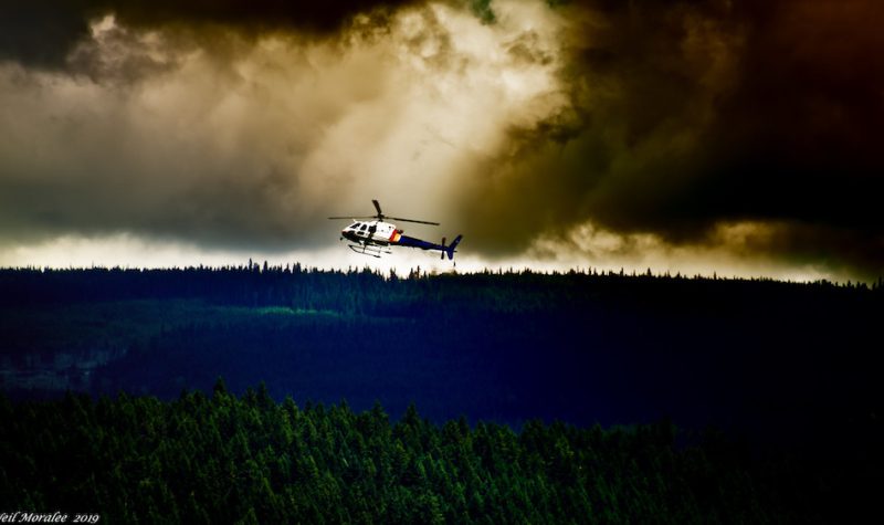 A helicopter flies above a forest with a lot of smoke billowing from the horizon
