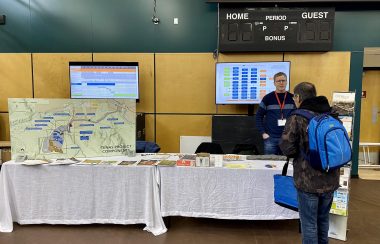 a man stands behind a table with a display map indicating the land where the proposed telkwa coal mine will go. Another man is standing in front of him reading information at a job fair.