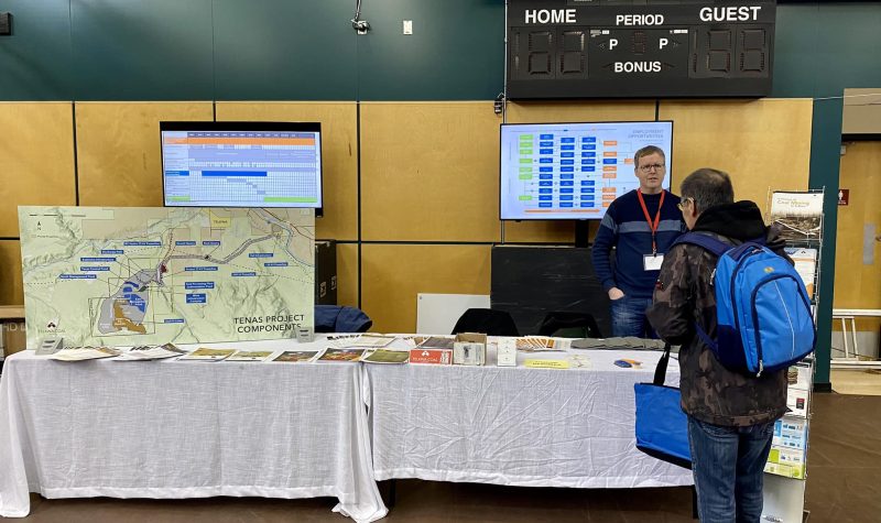a man stands behind a table with a display map indicating the land where the proposed telkwa coal mine will go. Another man is standing in front of him reading information at a job fair.
