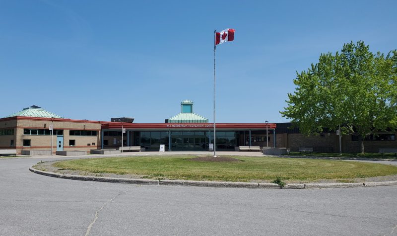 Large Recreation Facility spans left to right. Tree on the right side. A Canadian flag waves on a flagpole in front. There is grass and a parking lot in front of the flag.