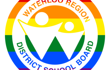 The circular Waterloo Region Distruct School Board Logo - that text on a white ring around 2 triangular people with their arms (?) stretched out. The logo is set on a pride rainbow background.